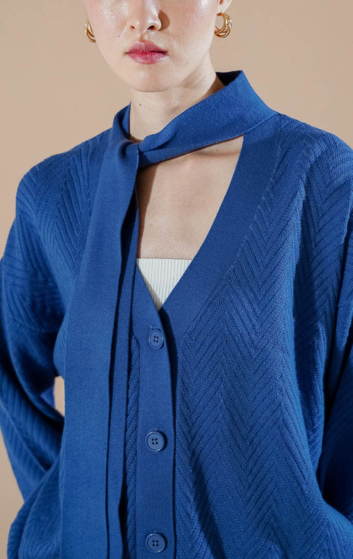 Marianne Cardigan Blue dari This Is April, cardigan rajut V-neck warna biru dengan kancing depan dan detail syal kain yang menyatu di bagian leher.