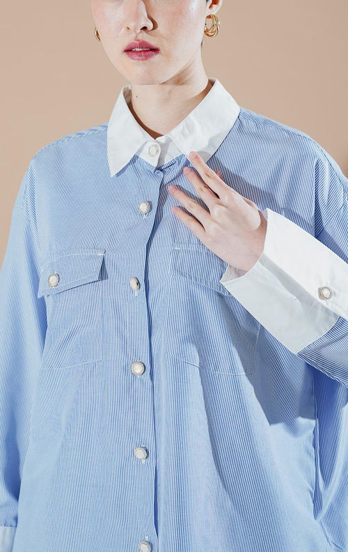 Model wanita mengenakan Merveille Top Blue Stripe dari This Is April, kemeja salur biru muda berpotongan longgar dengan kerah putih kontras dan dua saku depan.