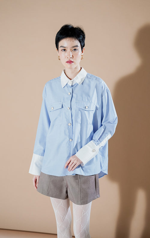 Model wanita mengenakan Merveille Top Blue Stripe dari This Is April, kemeja salur biru muda berpotongan longgar dengan kerah putih kontras dan dua saku depan.