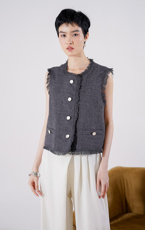 Rompi wanita tanpa lengan Mirabelle Vest dari This Is April berwarna abu-abu gelap (dark grey) dengan detail tepian raw edge yang menjuntai, dilengkapi kancing depan aktif dan aksen kancing saku, digantung di rak pakaian. 