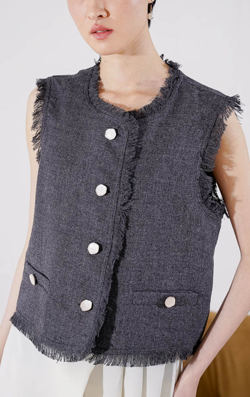 Rompi wanita tanpa lengan Mirabelle Vest dari This Is April berwarna abu-abu gelap (dark grey) dengan detail tepian raw edge yang menjuntai, dilengkapi kancing depan aktif dan aksen kancing saku, digantung di rak pakaian. 