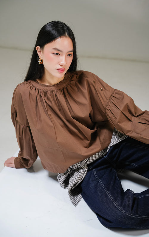 Blouse lengan panjang wanita This Is April Morgana Top warna cokelat tua (dark brown), model crewneck dengan detail kerutan penuh di sekeliling leher dan deretan kancing aktif di sisi bahu kiri. 