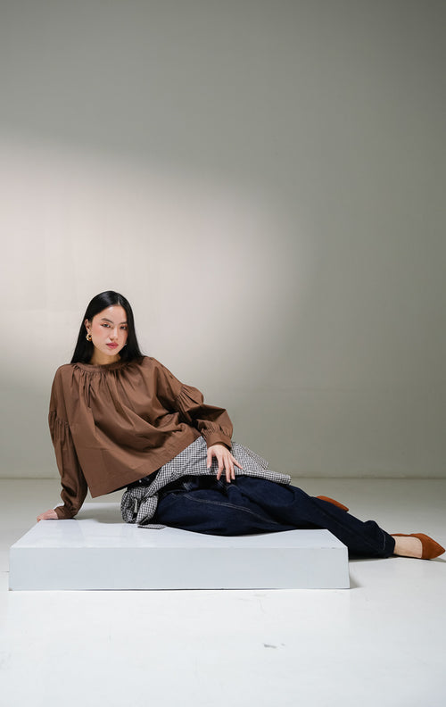 Blouse lengan panjang wanita This Is April Morgana Top warna cokelat tua (dark brown), model crewneck dengan detail kerutan penuh di sekeliling leher dan deretan kancing aktif di sisi bahu kiri. 