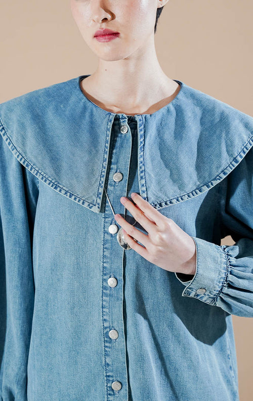 Poppi Top Denim dari This Is April, kemeja denim lengan panjang warna biru dengan detail kerah pelaut (sailor collar) yang lebar dan kancing depan perak.