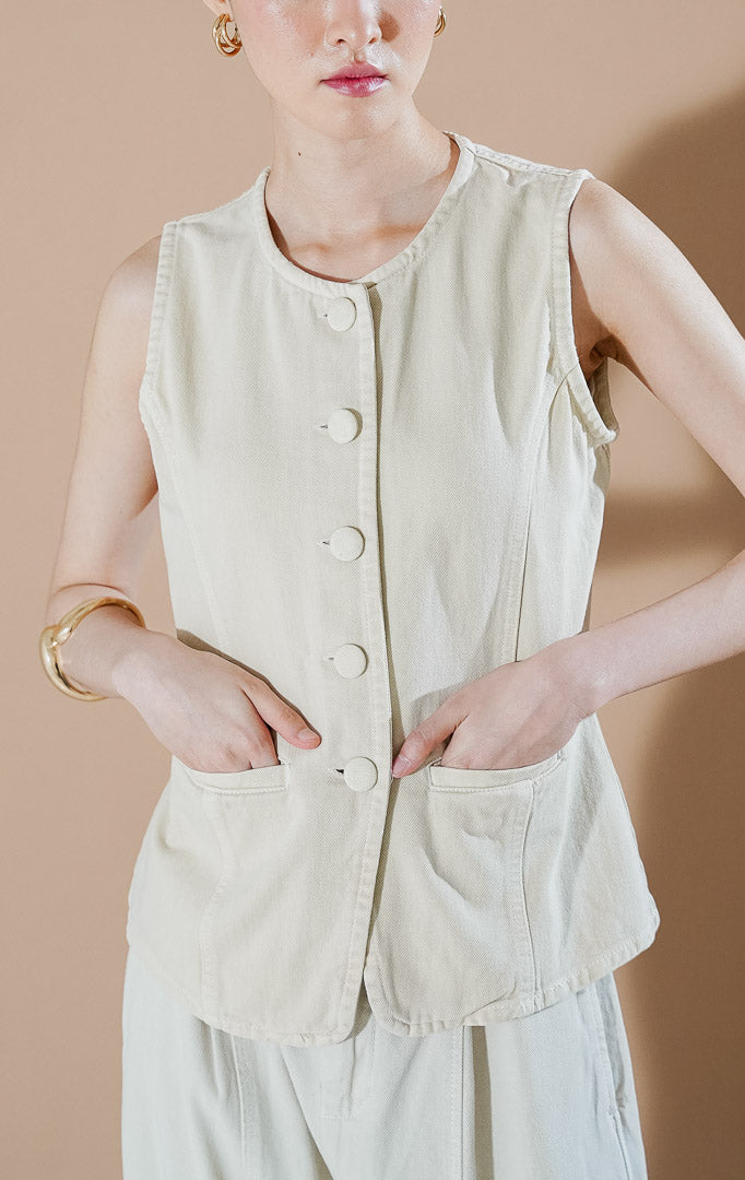 Rielle Vest Ivory dari This Is April, rompi denim tanpa lengan warna putih gading dengan kancing depan senada, kerah bulat, dan dua saku fungsional di bagian depan.