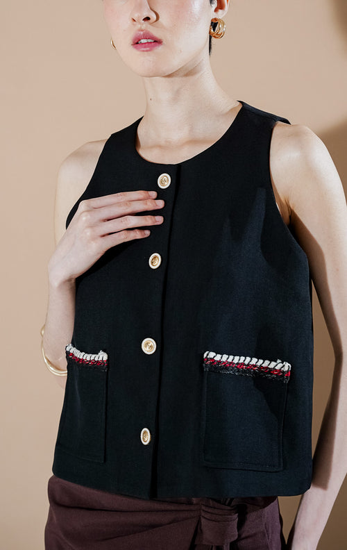 Model wanita mengenakan This Is April Shailene Vest Black, rompi hitam tanpa lengan dengan kancing depan, dipadukan dengan rok celana cokelat.