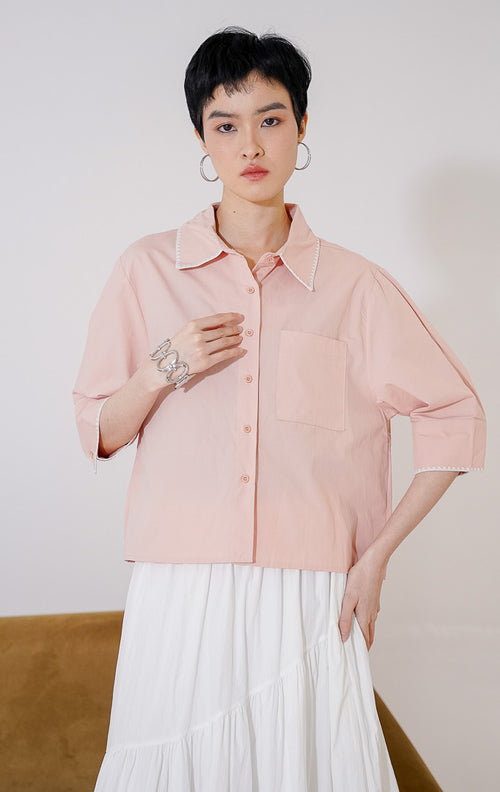 Kemeja wanita lengan 3/4 Venus Top dari This Is April berwarna dusty pink. Dilengkapi detail jahitan zigzag putih di tepi kerah dan ujung lengan, saku dada kiri fungsional, dan kancing depan, digantung di rak pakaian. 