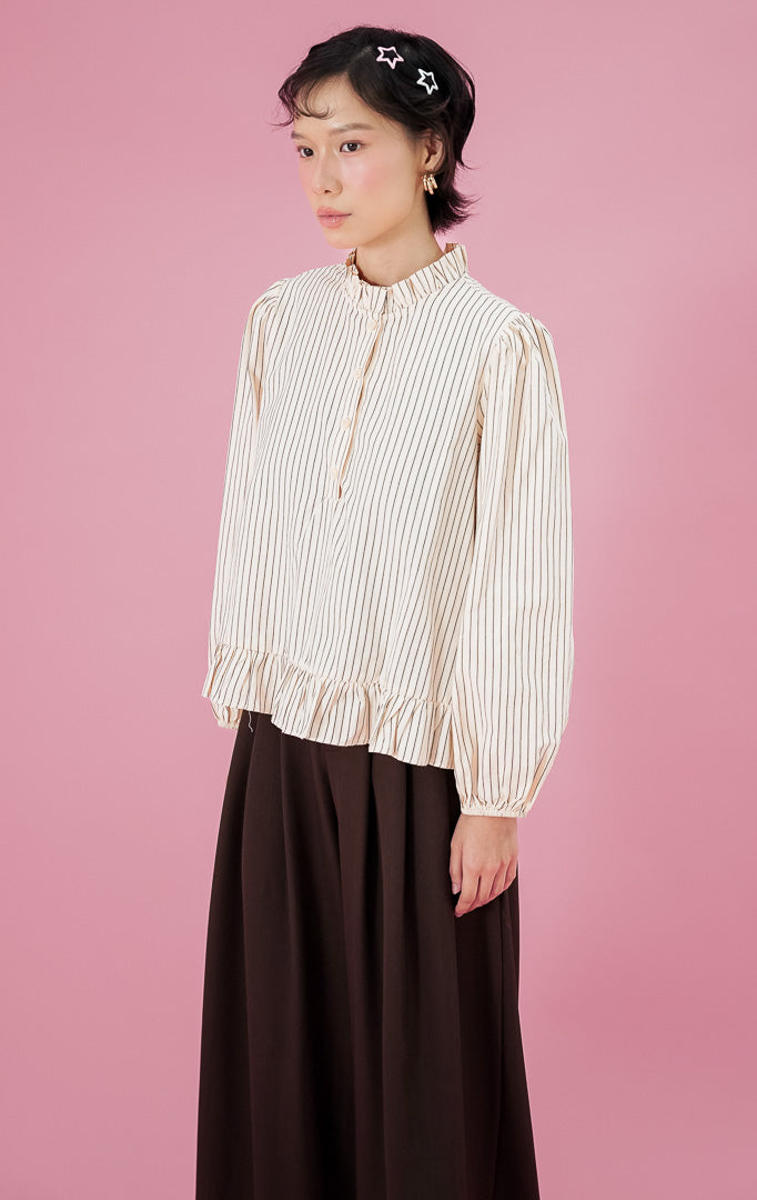 Model wanita mengenakan blouse lengan panjang This Is April Alessia Top warna butter yellow motif garis dengan detail frill di kerah dan bawah baju, dipadukan dengan celana kulot hitam lebar.