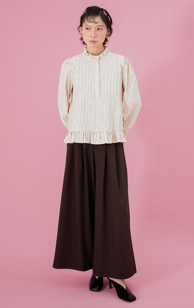 Model wanita mengenakan blouse lengan panjang This Is April Alessia Top warna butter yellow motif garis dengan detail frill di kerah dan bawah baju, dipadukan dengan celana kulot hitam lebar.