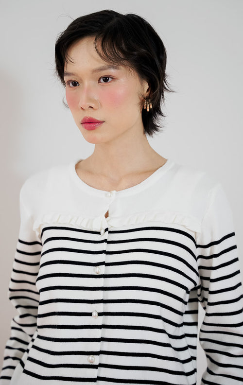 Model wanita mengenakan cardigan rajut lengan panjang This Is April Alexiz Cardigan warna putih motif garis hitam dengan kancing depan dan detail frill, dipadukan dengan celana panjang cokelat dan sepatu boots putih.