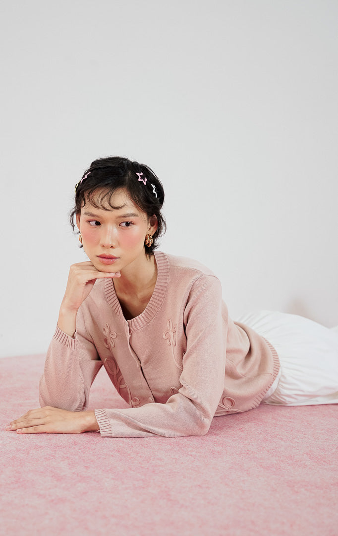 Model wanita mengenakan cardigan rajut lengan panjang This Is April Alistaire Cardigan warna dusty pink dengan kancing depan dan detail bordir bunga, dipadukan dengan rok midi putih dan sepatu flat hitam.