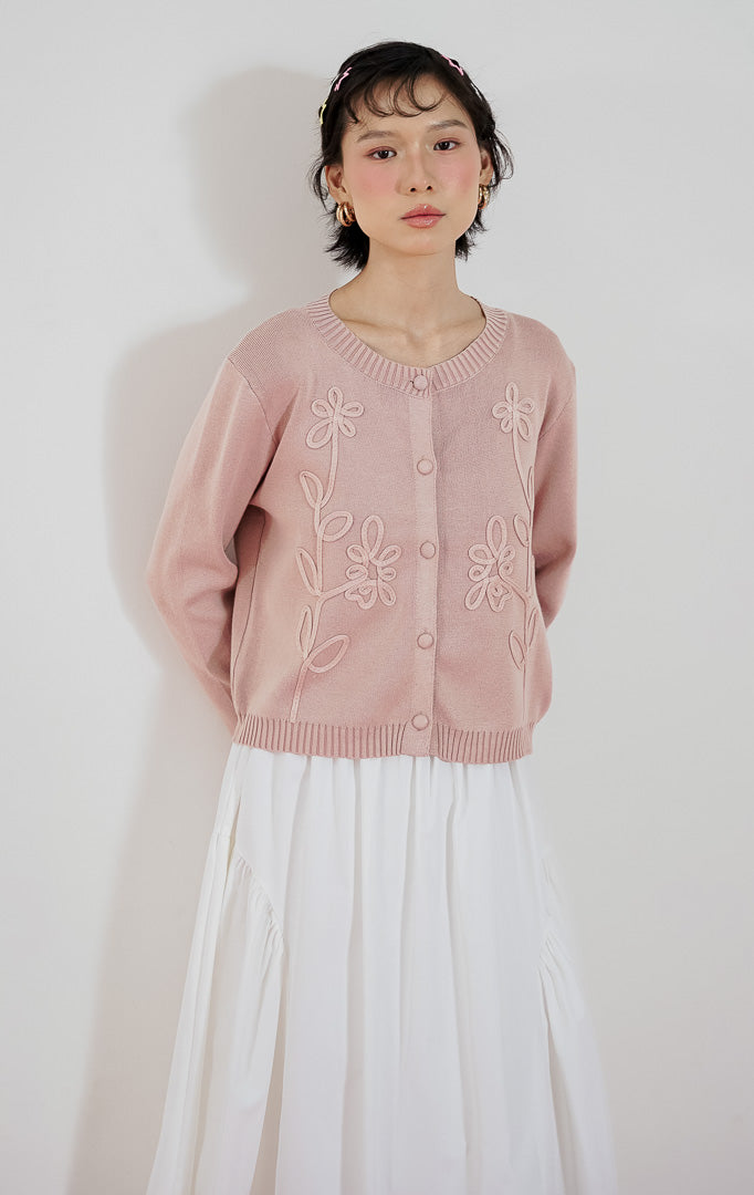 Model wanita mengenakan cardigan rajut lengan panjang This Is April Alistaire Cardigan warna dusty pink dengan kancing depan dan detail bordir bunga, dipadukan dengan rok midi putih dan sepatu flat hitam.