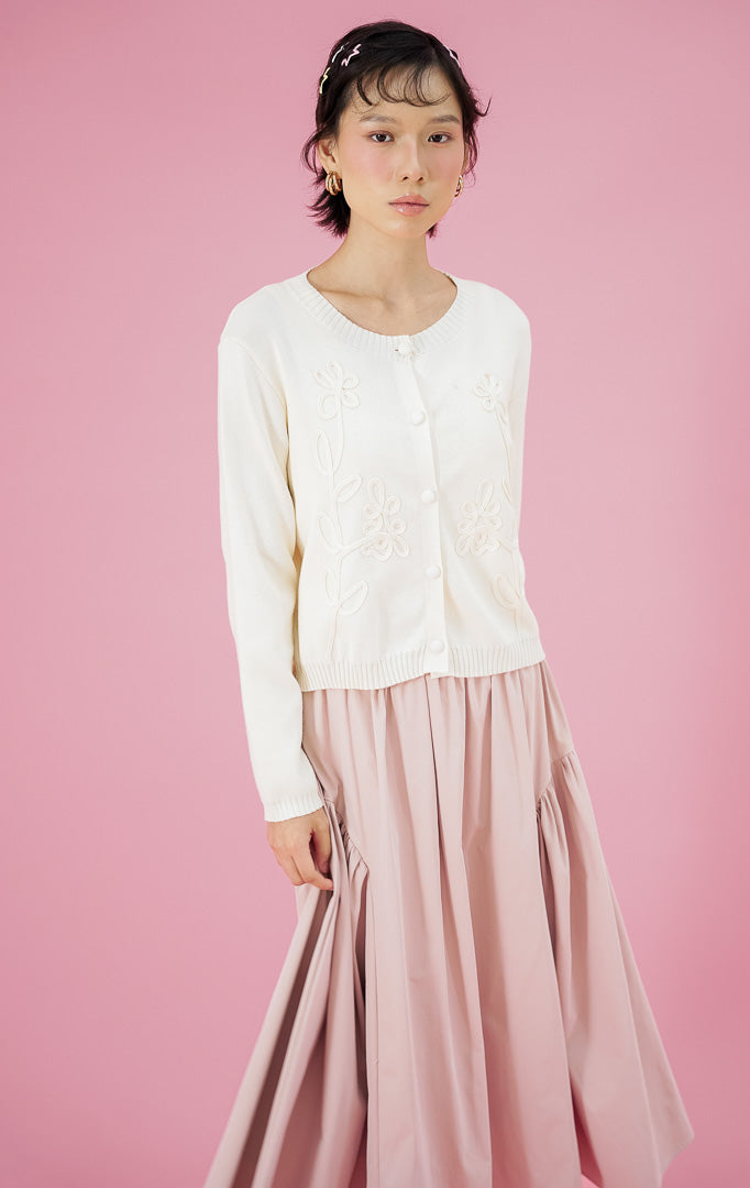 Model wanita mengenakan cardigan rajut lengan panjang This Is April Alistaire Cardigan warna ivory dengan kancing depan dan detail bordir bunga, dipadukan dengan rok midi lebar warna pink dan sepatu heels putih.