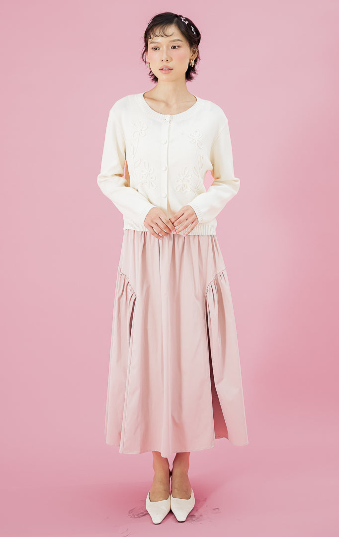 Model wanita mengenakan cardigan rajut lengan panjang This Is April Alistaire Cardigan warna ivory dengan kancing depan dan detail bordir bunga, dipadukan dengan rok midi lebar warna pink dan sepatu heels putih.