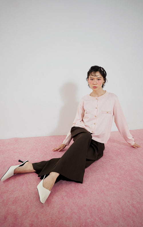 Model wanita mengenakan cardigan rajut klasik lengan panjang This Is April Arnon Cardigan warna pink muda dengan detail kancing emas, dipadukan dengan celana kulot lebar cokelat gelap dan sepatu heels putih.