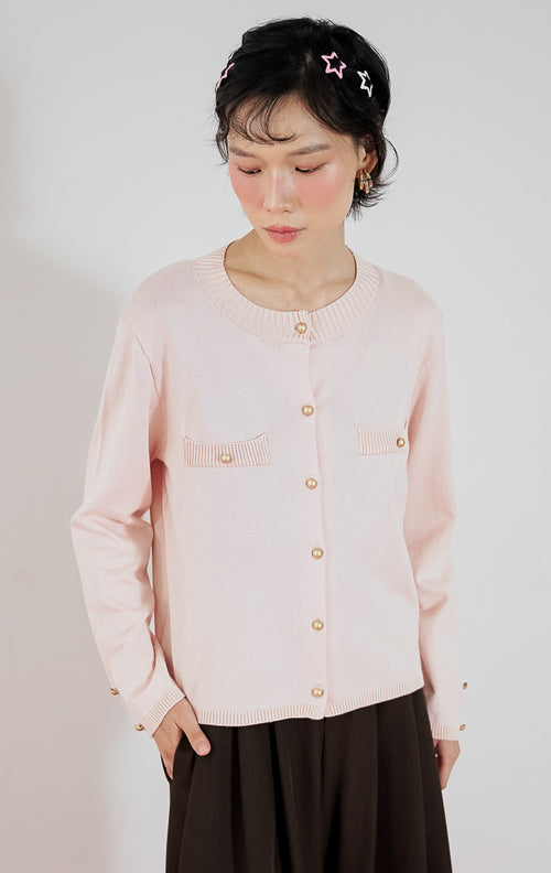 Model wanita mengenakan cardigan rajut klasik lengan panjang This Is April Arnon Cardigan warna pink muda dengan detail kancing emas, dipadukan dengan celana kulot lebar cokelat gelap dan sepatu heels putih.
