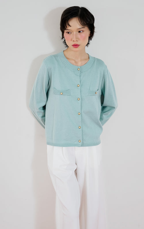 Model wanita mengenakan cardigan rajut klasik lengan panjang This Is April Arnon Cardigan warna tosca dengan kancing emas dan aksen saku, dipadukan dengan celana panjang kulot putih dan sepatu flat putih.