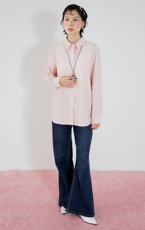 Model wanita mengenakan kemeja lengan panjang katun This Is April Athena Top warna light pink dengan kancing depan, dipadukan dengan celana jeans panjang biru tua, kalung aksesoris, dan sepatu putih.