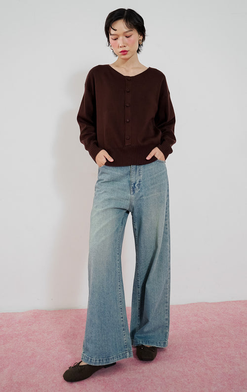 Model wanita mengenakan sweater rajut lengan panjang This Is April Avena Sweater warna cokelat dengan kancing depan, dipadukan dengan celana jeans panjang biru muda berpotongan lebar dan sepatu flat cokelat.