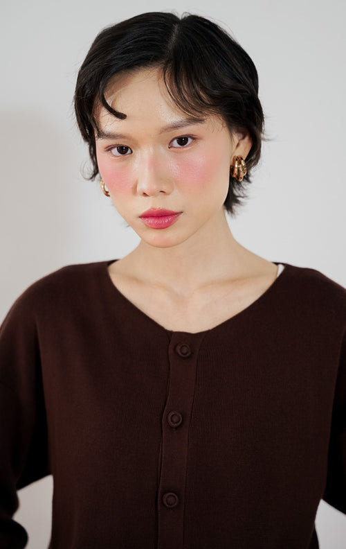 Model wanita mengenakan sweater rajut lengan panjang This Is April Avena Sweater warna cokelat dengan kancing depan, dipadukan dengan celana jeans panjang biru muda berpotongan lebar dan sepatu flat cokelat.