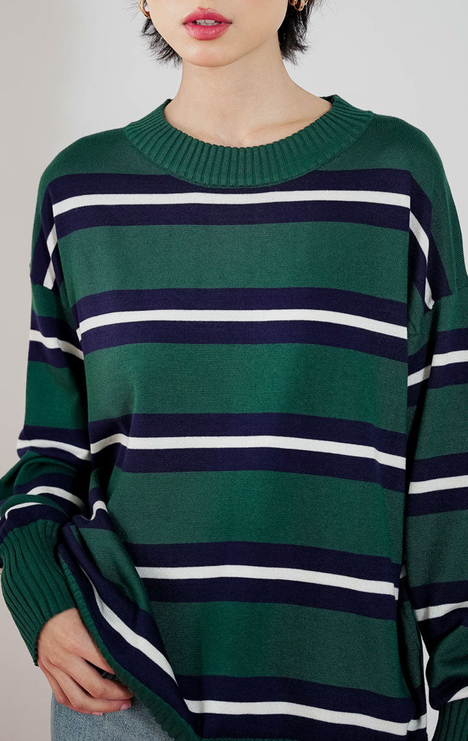 Model wanita mengenakan sweater rajut oversized This Is April Axton Sweater warna dark green motif garis putih, dipadukan dengan celana jeans wide-leg dan heels putih.