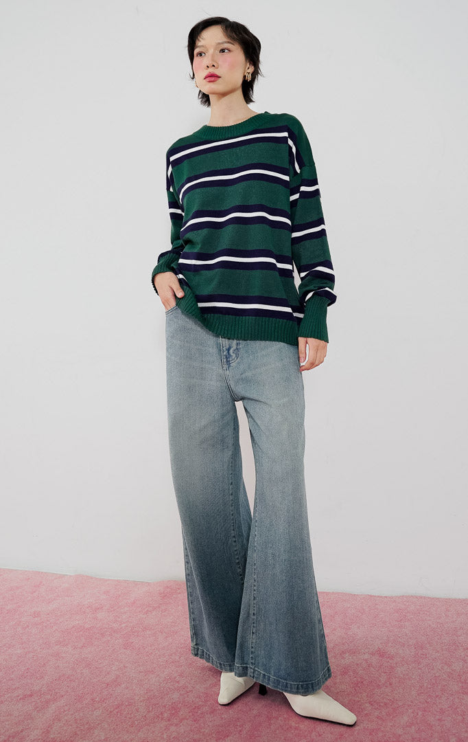Model wanita mengenakan sweater rajut oversized This Is April Axton Sweater warna dark green motif garis putih, dipadukan dengan celana jeans wide-leg dan heels putih.