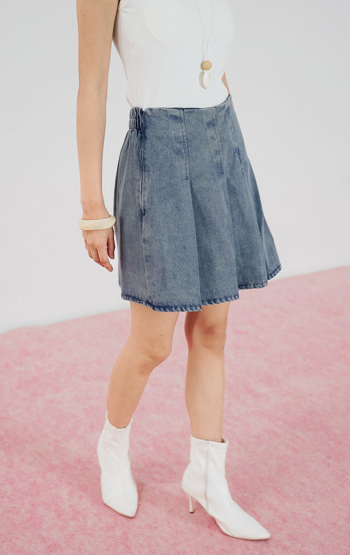 Model wanita mengenakan rok celana denim pendek This Is April Calypso Skort warna light denim dengan detail pleat depan, dipadukan dengan tanktop putih dan sepatu boots putih.