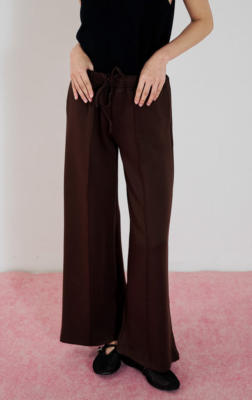 Model wanita mengenakan celana panjang This Is April Cora Pants warna dark brown berpotongan straight-leg dengan detail tali serut di pinggang, dipadukan dengan tanktop hitam.