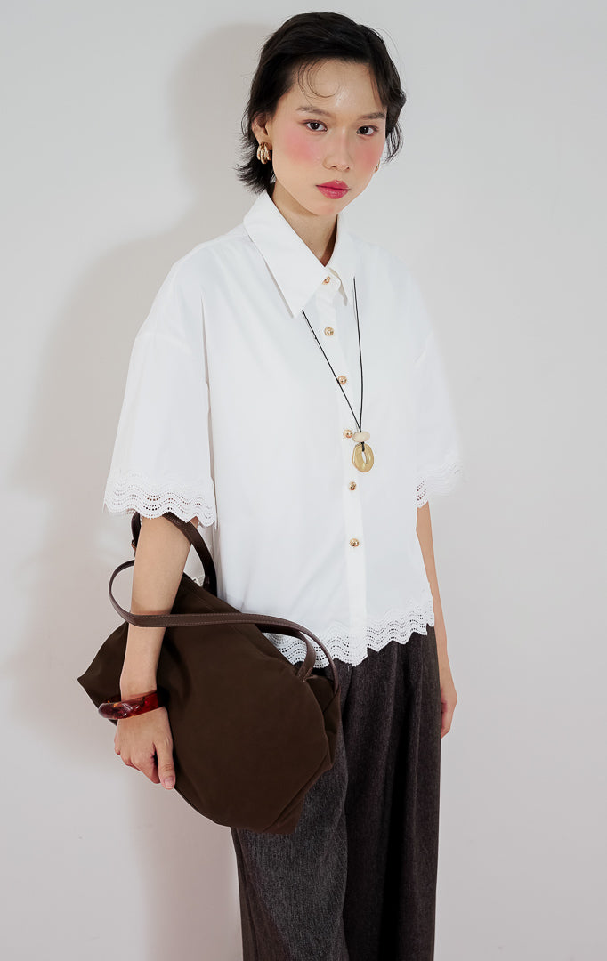 Model wanita mengenakan atasan lengan pendek kemeja putih This Is April Corbin Top dengan detail renda crochet di ujung lengan dan bawah baju, dipadukan dengan celana panjang cokelat.