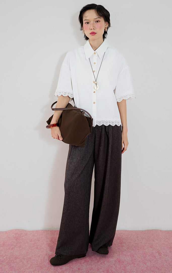 Model wanita mengenakan atasan lengan pendek kemeja putih This Is April Corbin Top dengan detail renda crochet di ujung lengan dan bawah baju, dipadukan dengan celana panjang cokelat.