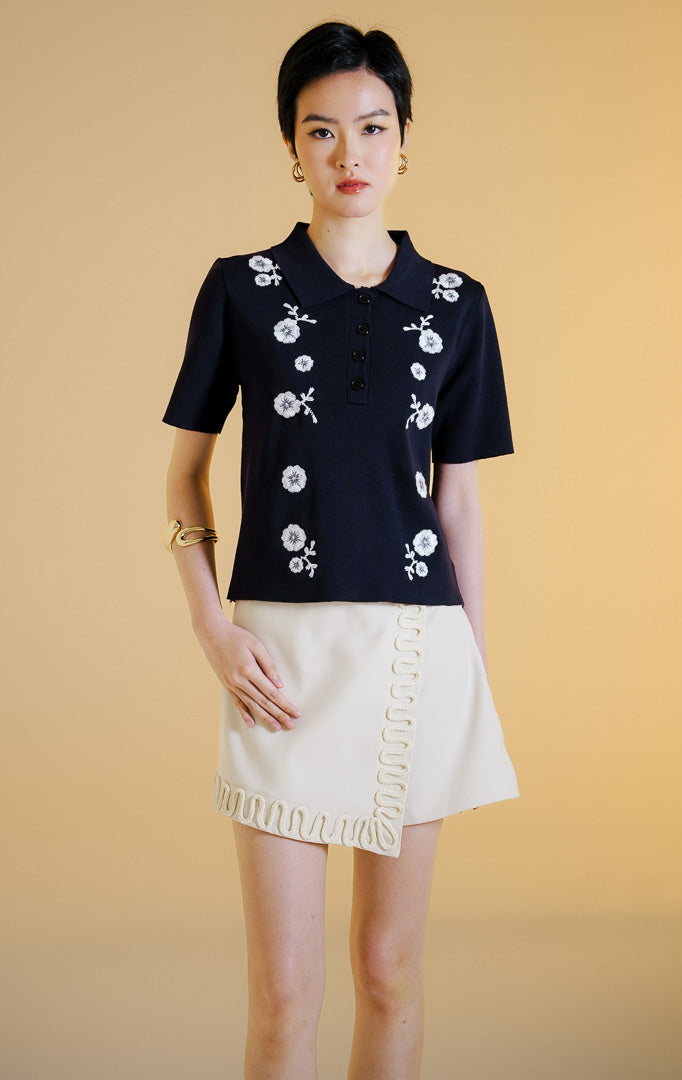 Pakaian wanita model atasan polo rajut lengan pendek berkerah dengan kancing depan dan detail bordir bunga (floral embroidered knit polo top) warna navy Elenora Top untuk gaya kasual anggun. 