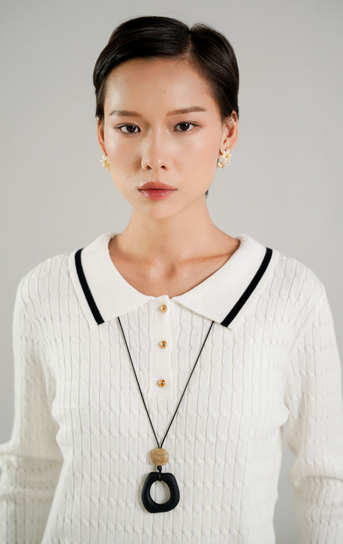 Sweater rajut kabel (cable knit) wanita lengan panjang warna putih dengan kerah polo, detail tiga kancing emas di dada, serta aksen garis hitam di kerah dan ujung baju dari This Is April. 