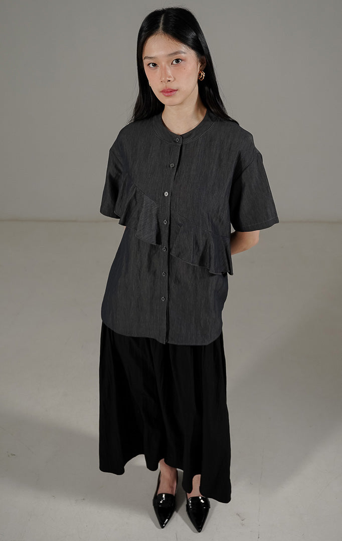 Karina Top Dark Grey dari This Is April, kemeja lengan pendek wanita warna abu abu tua dengan detail kerah bulat dan aksen ruffle asimetris di bagian depan.