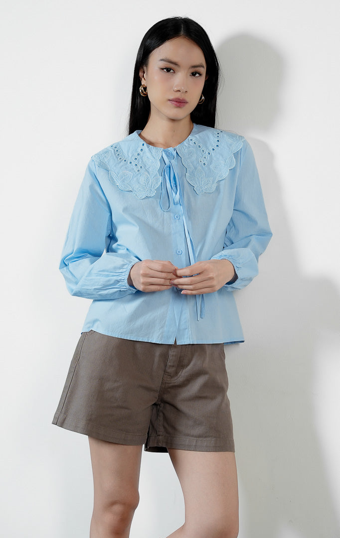 Pakaian wanita model blouse lengan panjang bahan katun dengan detail kerah bordir motif lubang (eyelet broderie) dan tepi gelombang (scalloped edges) warna biru muda Lioren Top untuk gaya kasual anggun. 