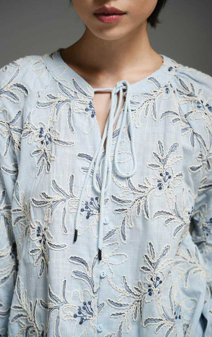 Blus wanita gaya bohemian lengan panjang warna biru muda (light blue bohemian floral embroidered top) Robena Top dengan aksen bordir motif bunga, bahan katun bertekstur, serta kancing dan tali serut depan yang elegan. 