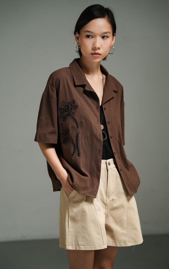 Kemeja wanita lengan pendek bahan linen warna cokelat tua (dark brown floral crochet shirt top) Ziven Top dengan detail rajut motif bunga yang rumit, penutup kancing depan, serta gaya kasual yang elegan. 