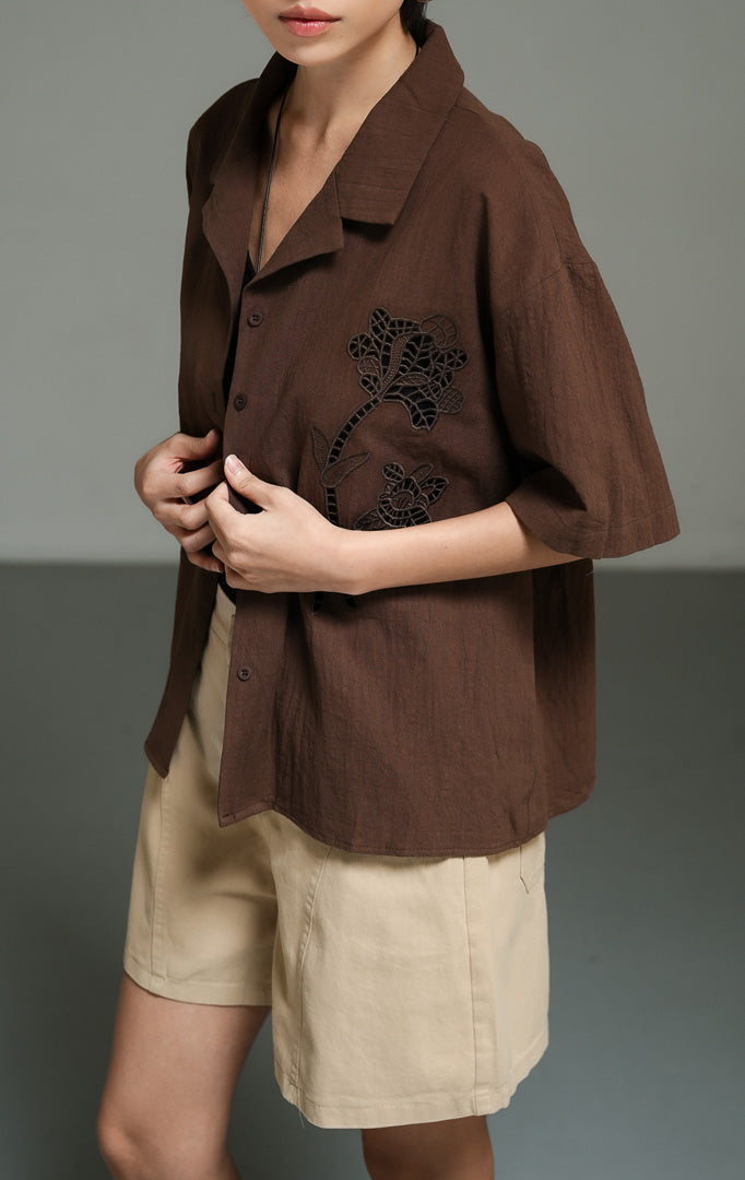 Kemeja wanita lengan pendek bahan linen warna cokelat tua (dark brown floral crochet shirt top) Ziven Top dengan detail rajut motif bunga yang rumit, penutup kancing depan, serta gaya kasual yang elegan. 