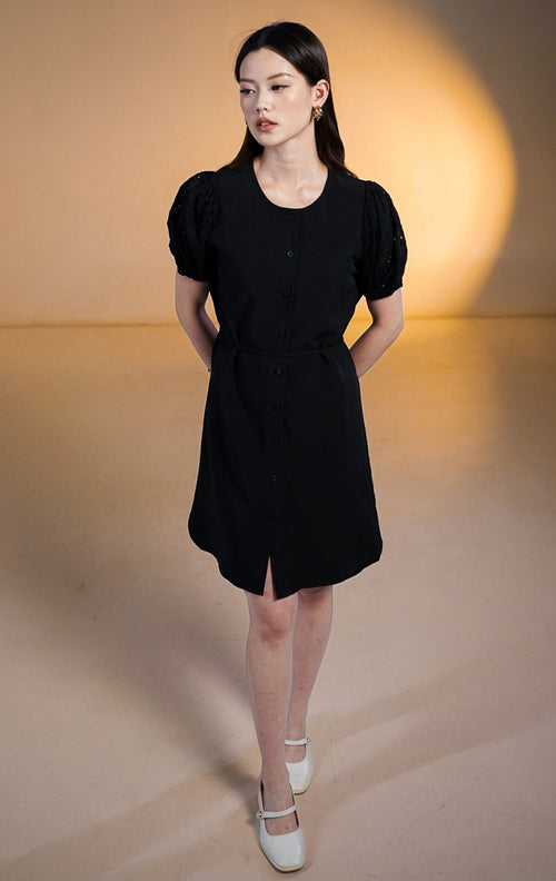 Model wanita memakai Titania Dress Black, gaun mini kancing depan warna hitam dengan lengan puff detail bordir eyelet dan tali pinggang