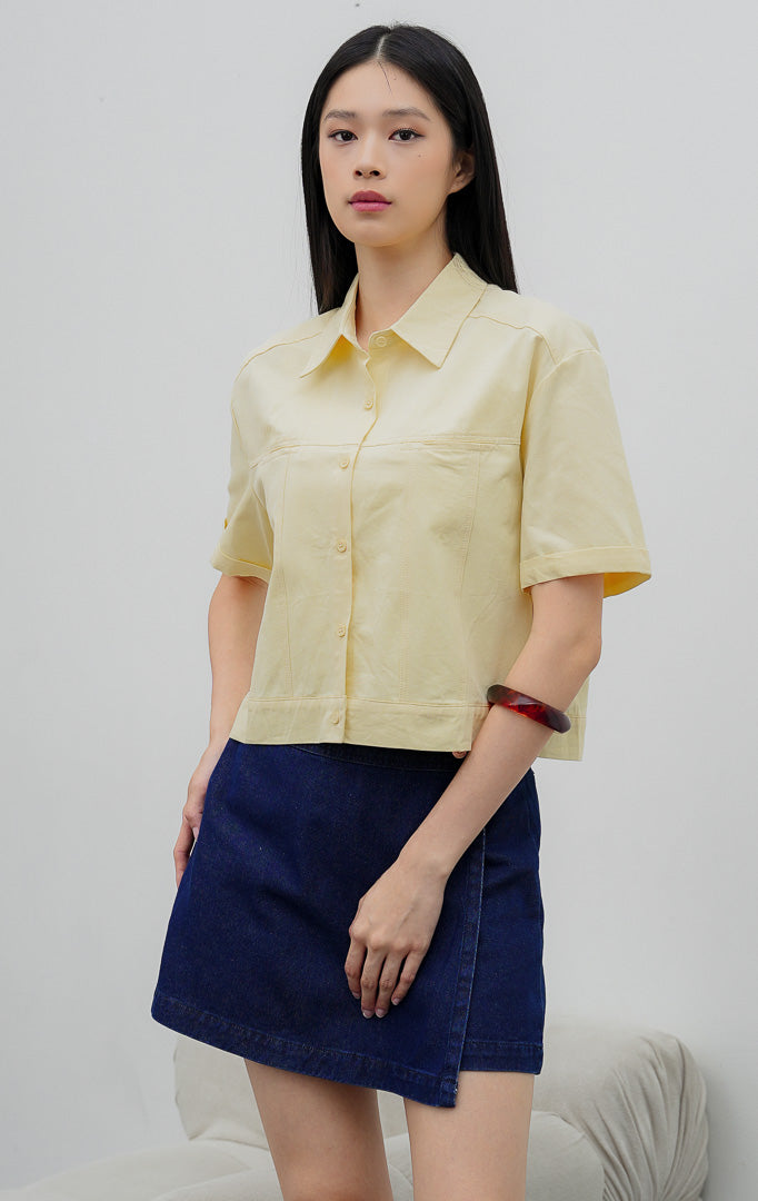 Kemeja kancing depan wanita warna kuning lembut (butter yellow) Velira Top berbahan katun terstruktur (structured cotton) dengan detail jahitan bahu (yoke seam detailing) dan lengan pendek. 