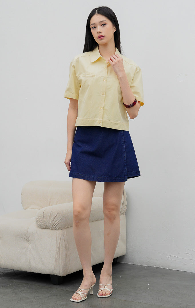 Kemeja kancing depan wanita warna kuning lembut (butter yellow) Velira Top berbahan katun terstruktur (structured cotton) dengan detail jahitan bahu (yoke seam detailing) dan lengan pendek. 