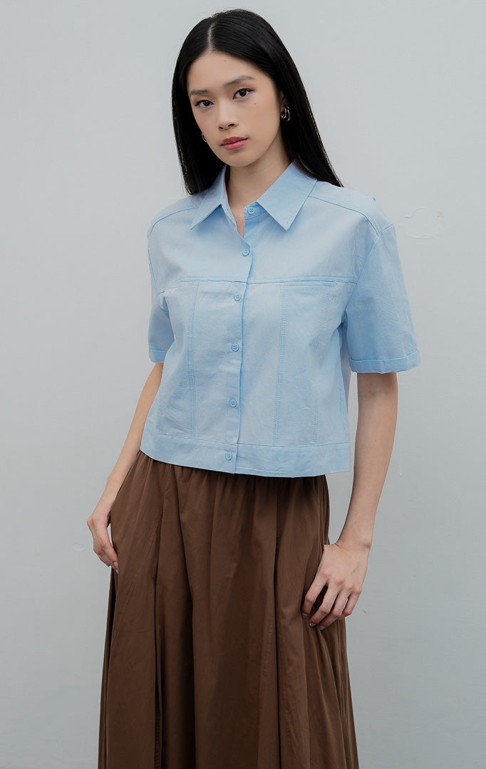 Kemeja kancing depan wanita warna biru muda (light blue) Velira Top berbahan katun terstruktur (structured cotton) dengan detail jahitan bahu (yoke seam detailing) dan lengan pendek.