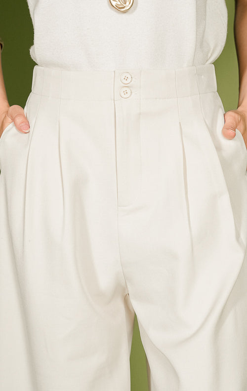 Celana panjang wanita Vincent Pants berpotongan lurus (straight cut) warna ivory atau krem terang, dilengkapi detail dua kancing bersusun (double button) di ban pinggang depan, aksen lipatan vertikal (pleats), saku samping, dan karet elastis di pinggang bagian belakang.