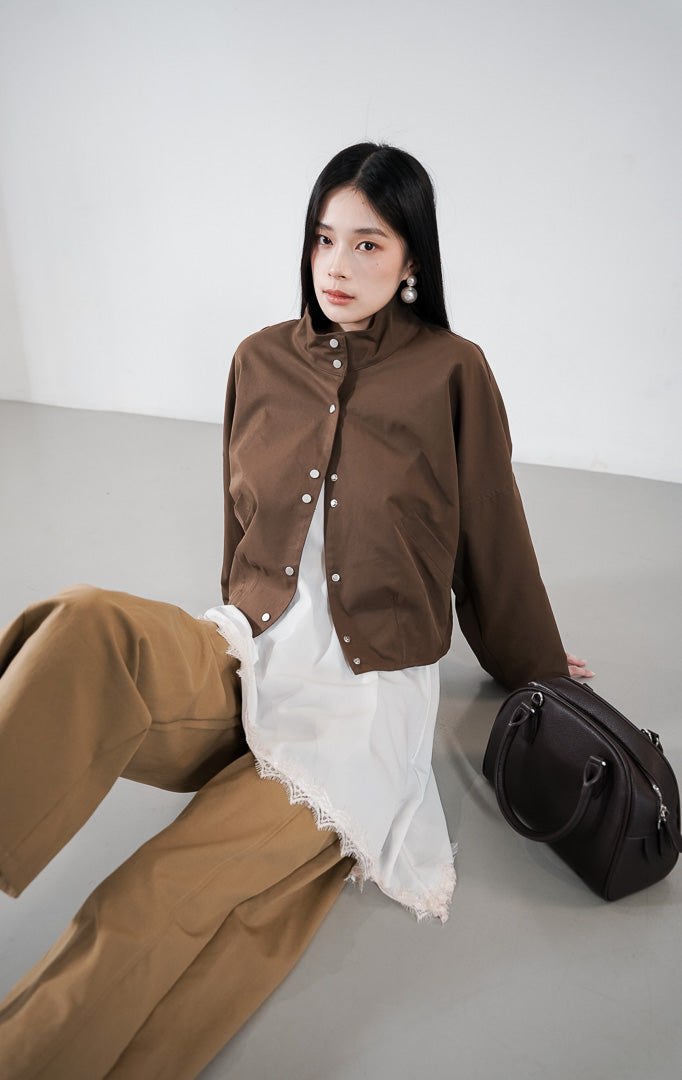 Vincenza Jacket warna dark brown, jaket lengan panjang dengan kerah tinggi (stand collar) dan deretan kancing perak, dipadukan dengan atasan putih berenda.