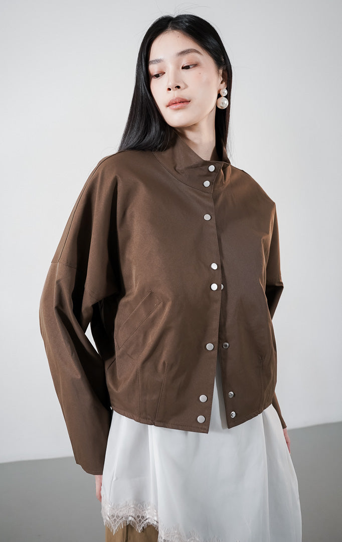Vincenza Jacket warna dark brown, jaket lengan panjang dengan kerah tinggi (stand collar) dan deretan kancing perak, dipadukan dengan atasan putih berenda1