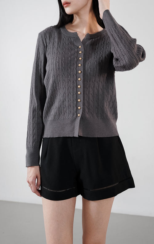 Vinnie Cardigan warna dark grey, kardigan rajut kabel (cable knit) lengan panjang dengan deretan kancing elegan, dipadukan dengan celana pendek hitam.