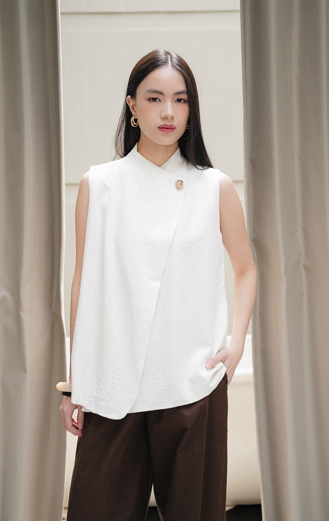 Rompi vest wanita tanpa lengan Viridiana Vest warna ivory krem dengan potongan siluet asimetris wrap berkerah tinggi dan aksen kancing emas elegan.