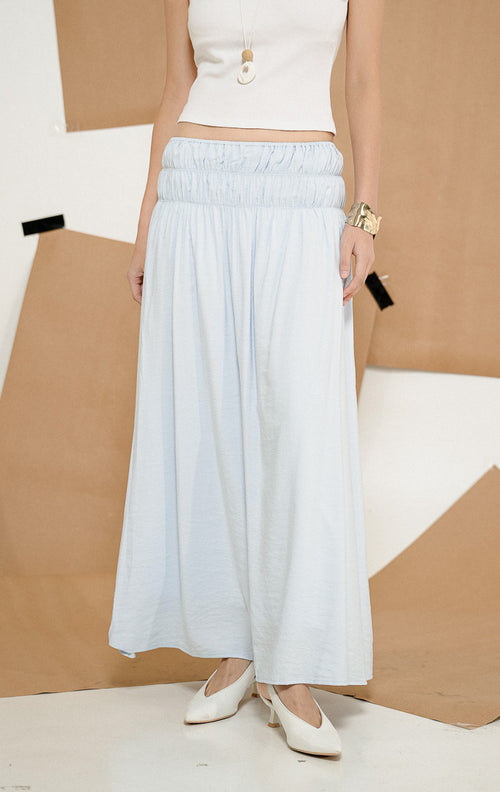 Model berdiri mengenakan Wattson Flowy Ruched Maxi Skirt Light Blue, produk rok panjang wanita model maxi warna biru muda dengan detail pinggang elastis berkerut lebar. 