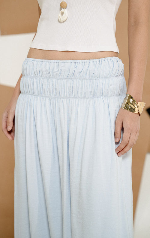 Model berdiri mengenakan Wattson Flowy Ruched Maxi Skirt Light Blue, produk rok panjang wanita model maxi warna biru muda dengan detail pinggang elastis berkerut lebar. 