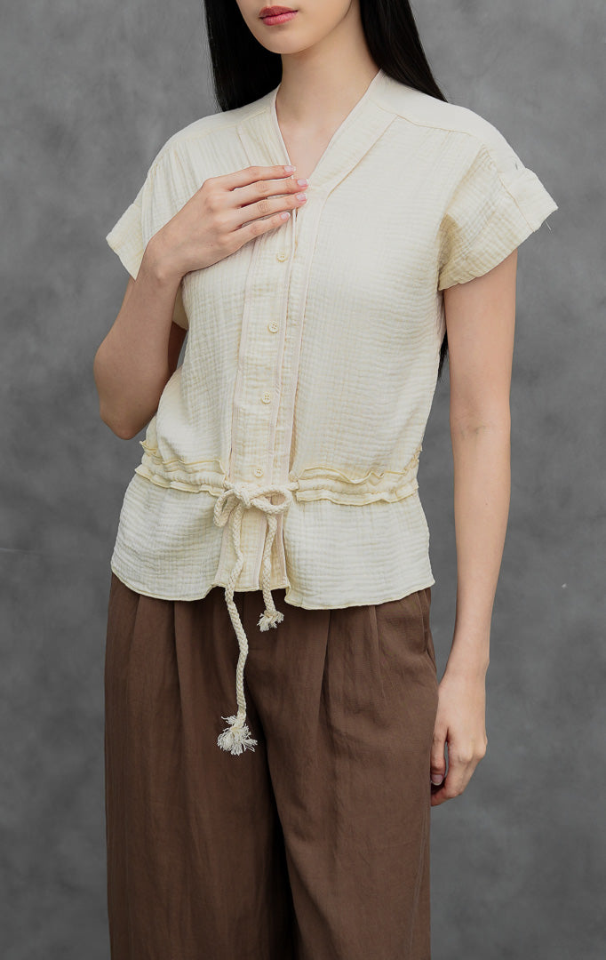 Blus wanita lengan pendek warna gading (ivory) Weasley Top berbahan katun bertekstur (textured cotton) dengan kerah V, bukaan kancing depan, tali serut pinggang (drawstring waist), dan detail lipatan kerut (ruffled hem).