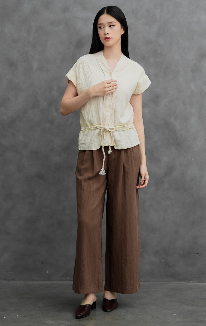 Blus wanita lengan pendek warna gading (ivory) Weasley Top berbahan katun bertekstur (textured cotton) dengan kerah V, bukaan kancing depan, tali serut pinggang (drawstring waist), dan detail lipatan kerut (ruffled hem).